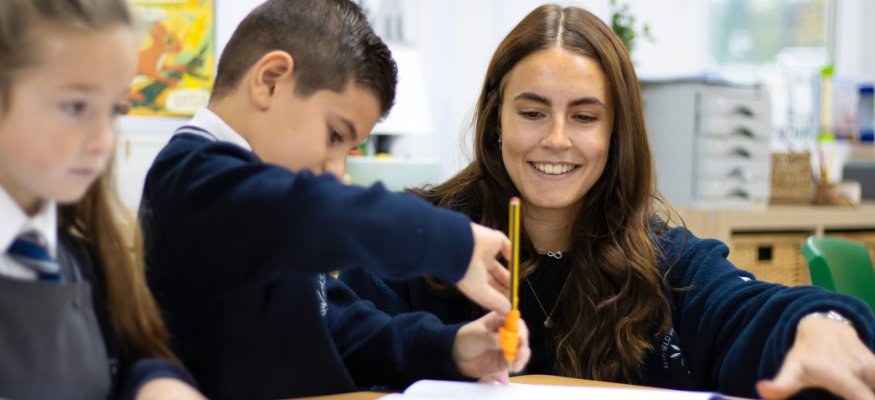 An apprentice in the classroom is smiling whilst supporting a primary aged child to complete their work.